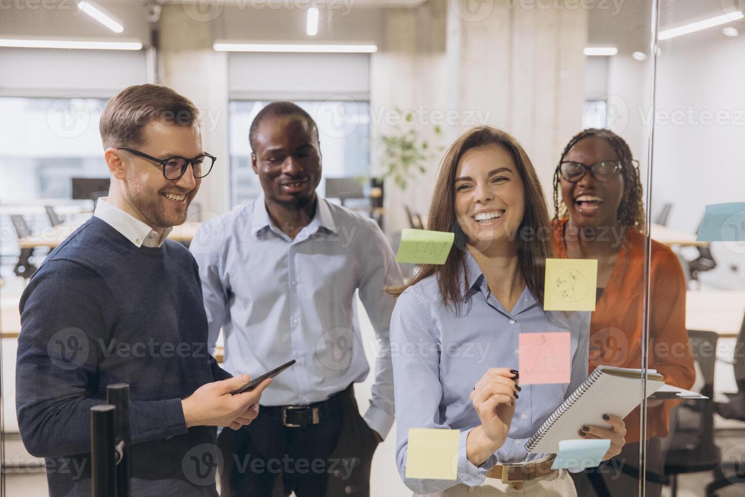 Diverse business team collaborates in a modern office, brainstorming ideas with sticky notes on glass. Smiling professionals engaged in creative discussion photo