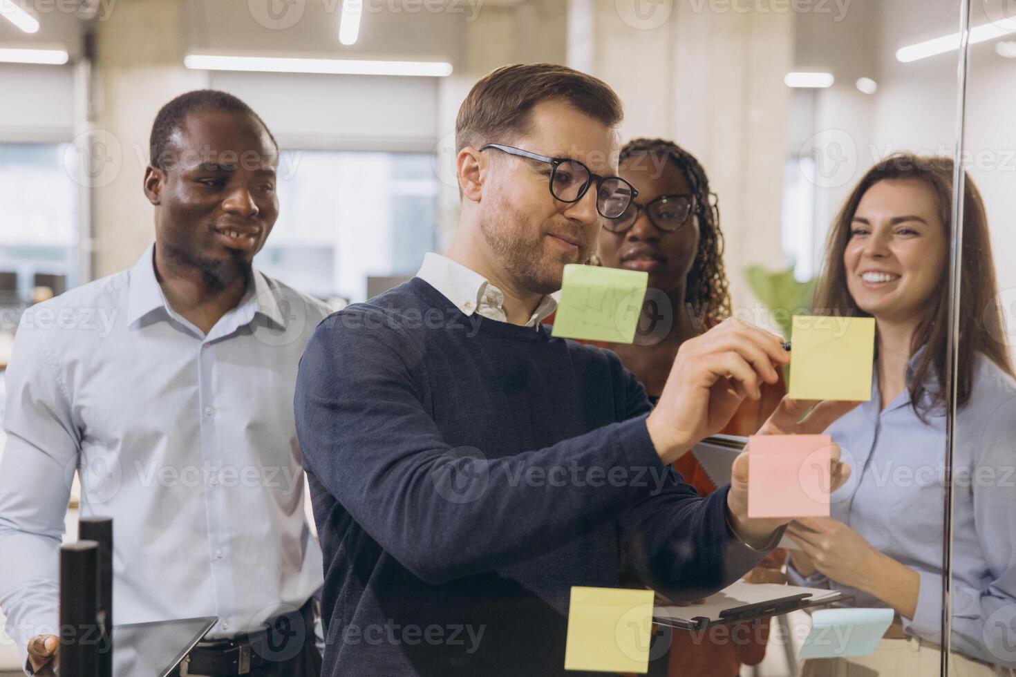 Diverse group of professionals engaging in a collaborative brainstorming session, using sticky notes on a glass wall in a modern office environment photo