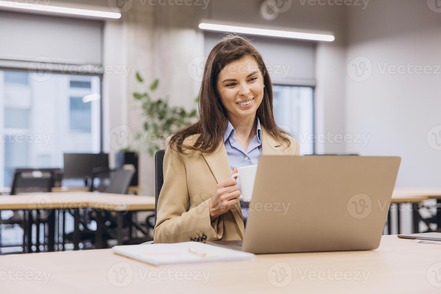 Smiling businesswoman enjoying a cup of coffee while working on a laptop in a bright, modern office space filled with natural light photo