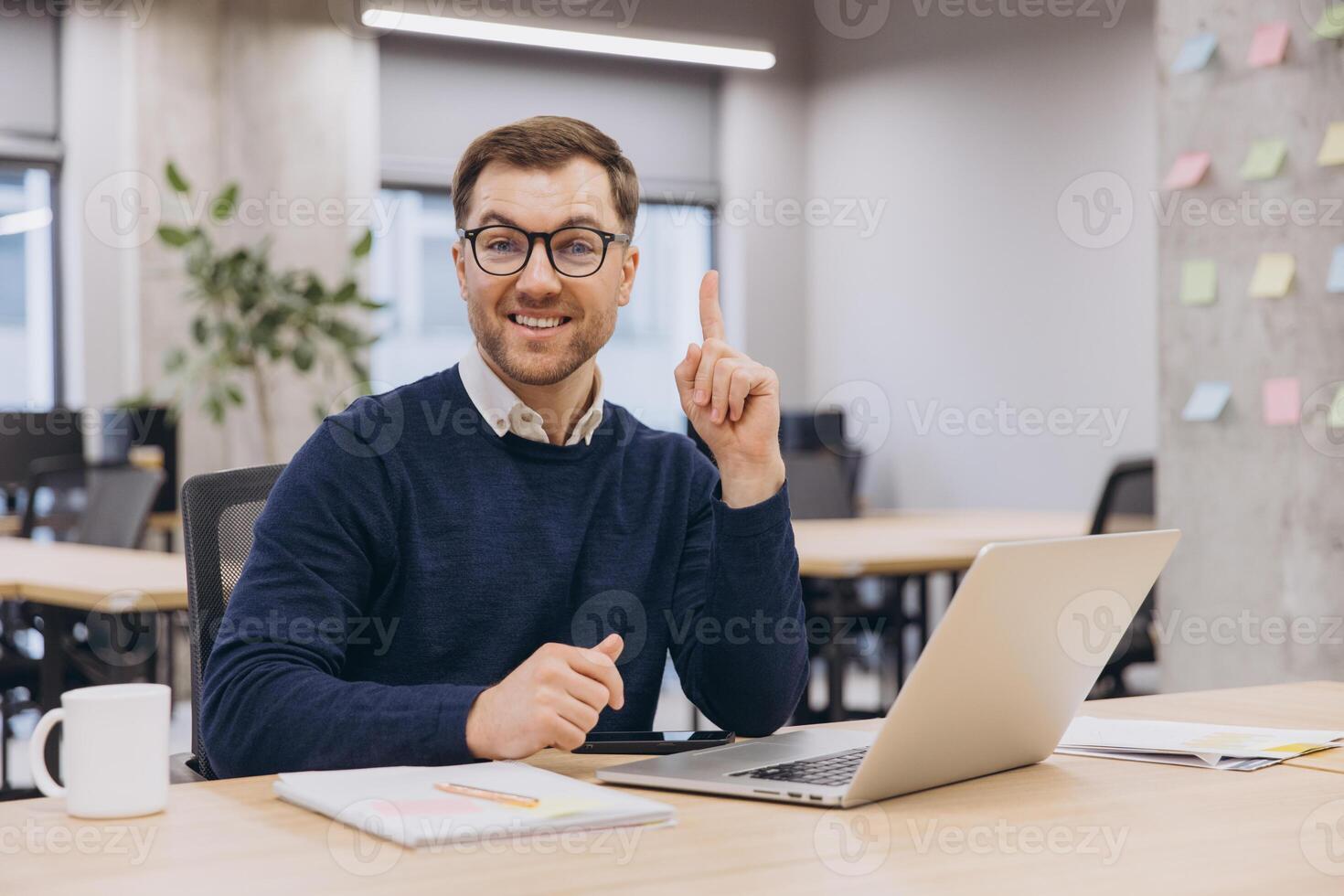 Smiling businessman raising his index finger having a good idea while working on his laptop in a modern office photo
