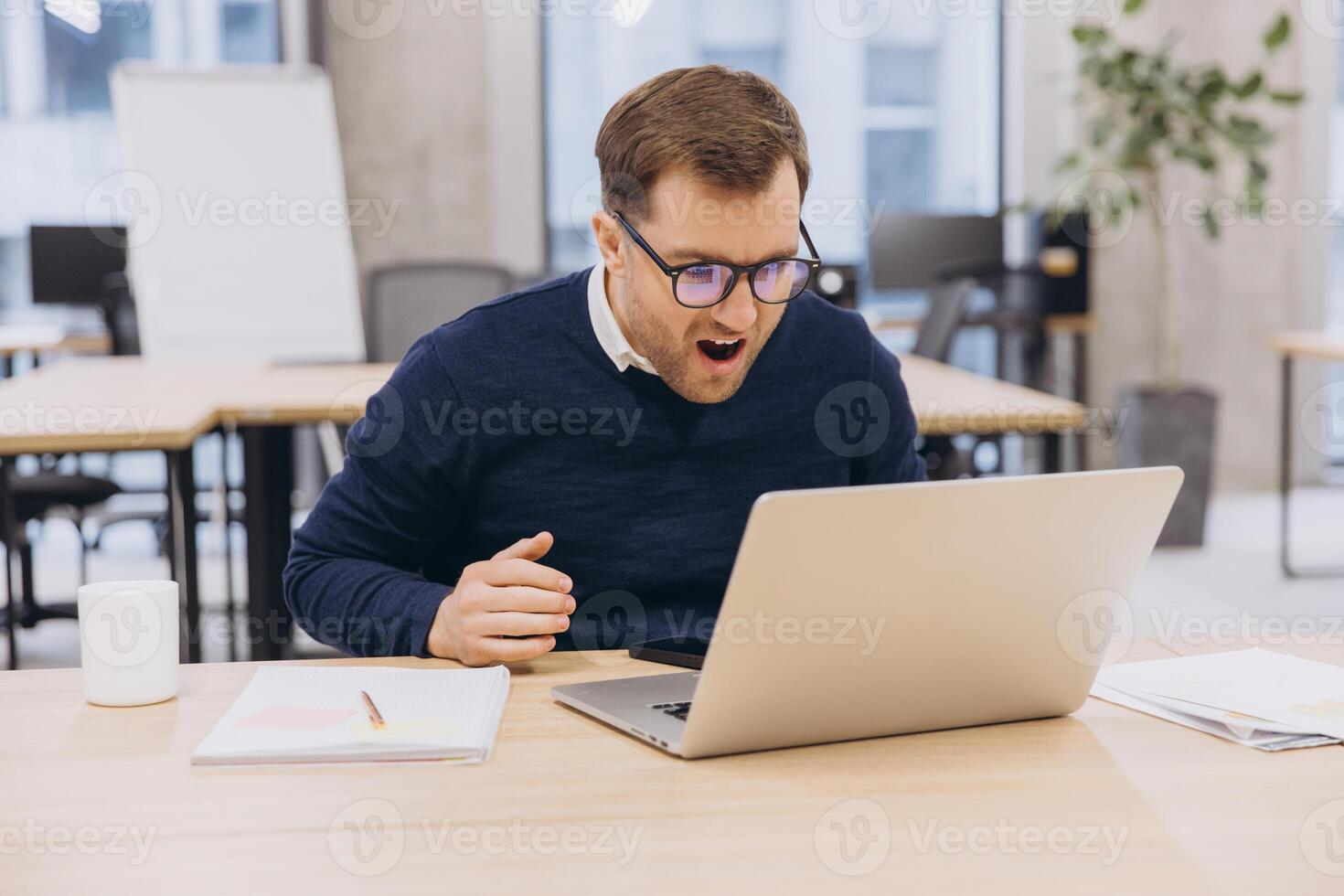 Businessman with glasses expressing surprise while working on his laptop at a modern office desk, surrounded by documents and a bright window photo