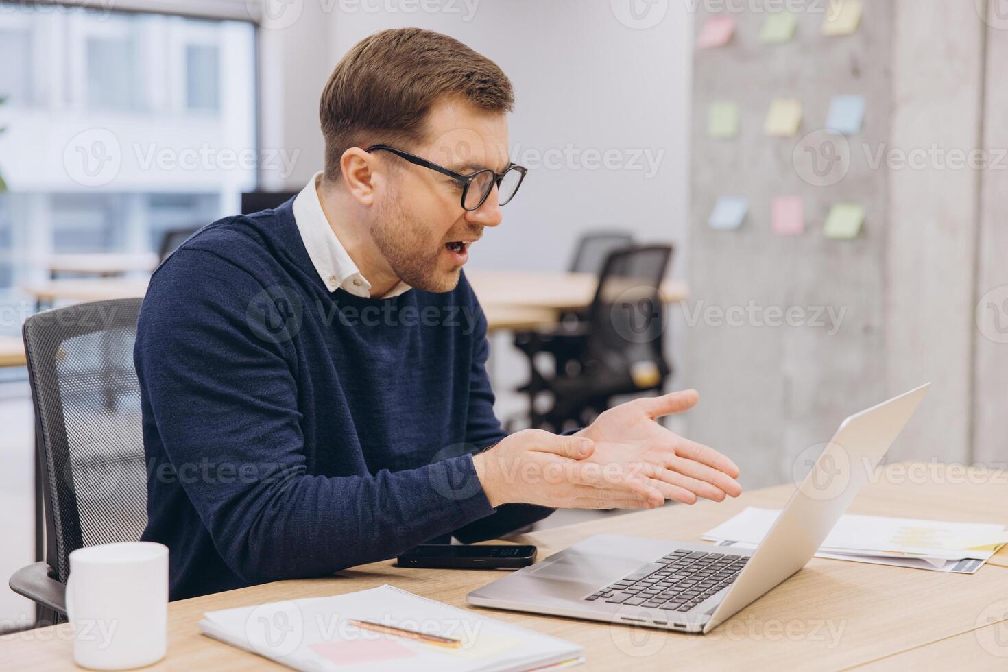 Professional businessman working remotely, gesturing during conference, using laptop at minimalist workspace with sleek design photo