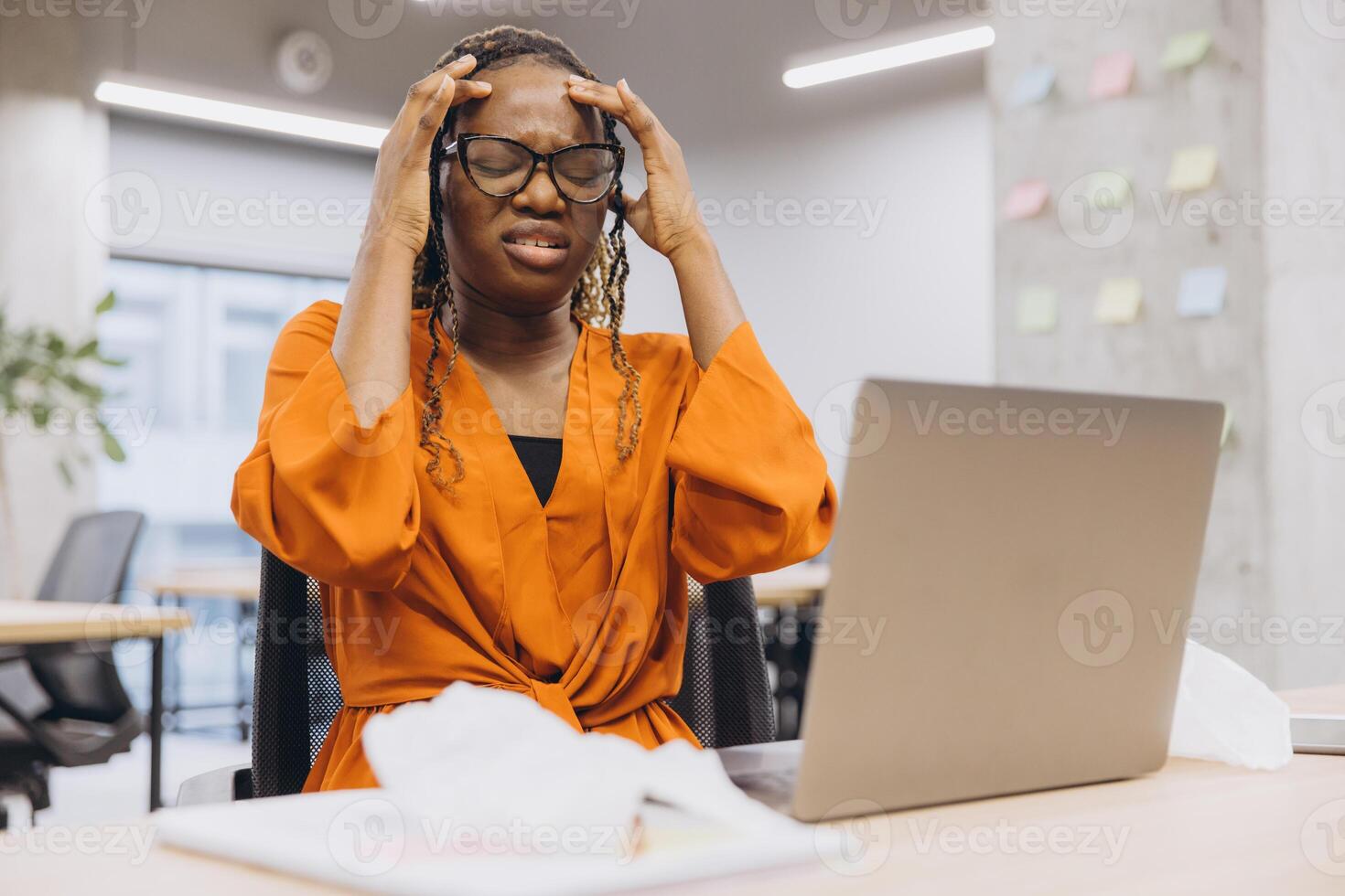 African american businesswoman experiencing stress while working on a laptop in a modern office setting, surrounded by paperwork and sticky notes photo