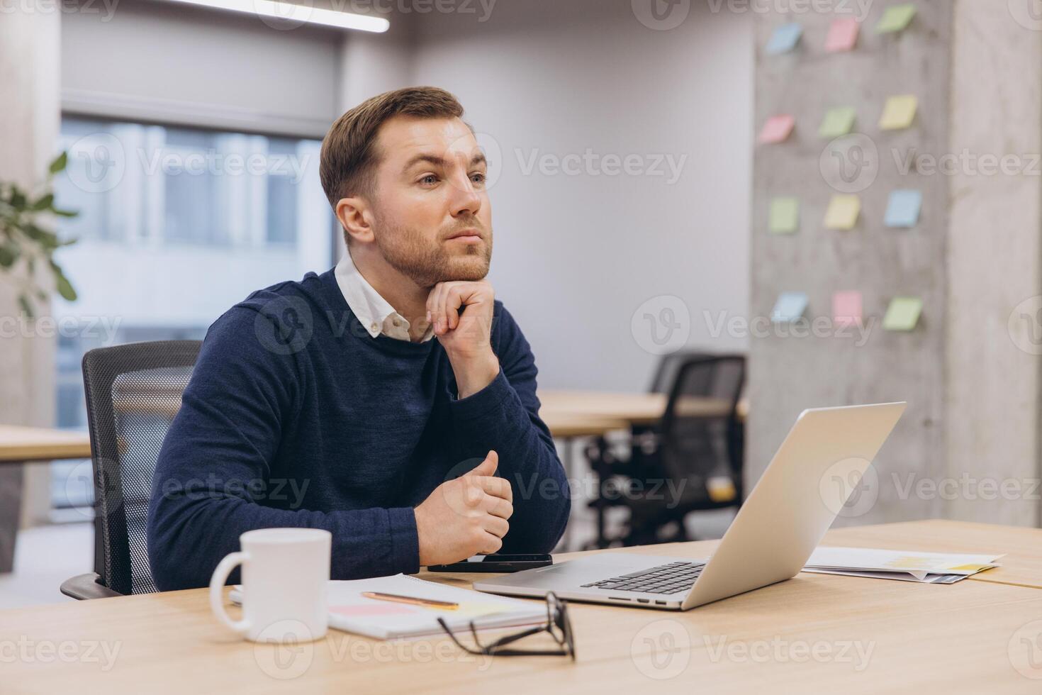 Professional businessman sitting at desk, resting chin on hand, gazing pensively while deep in strategic thought during workday photo