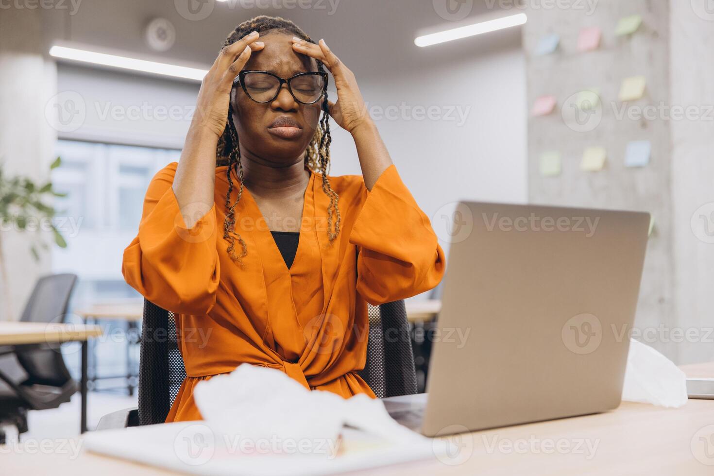 African American businesswoman in an office setting, holding her head in frustration while working on a laptop, surrounded by notes and papers photo