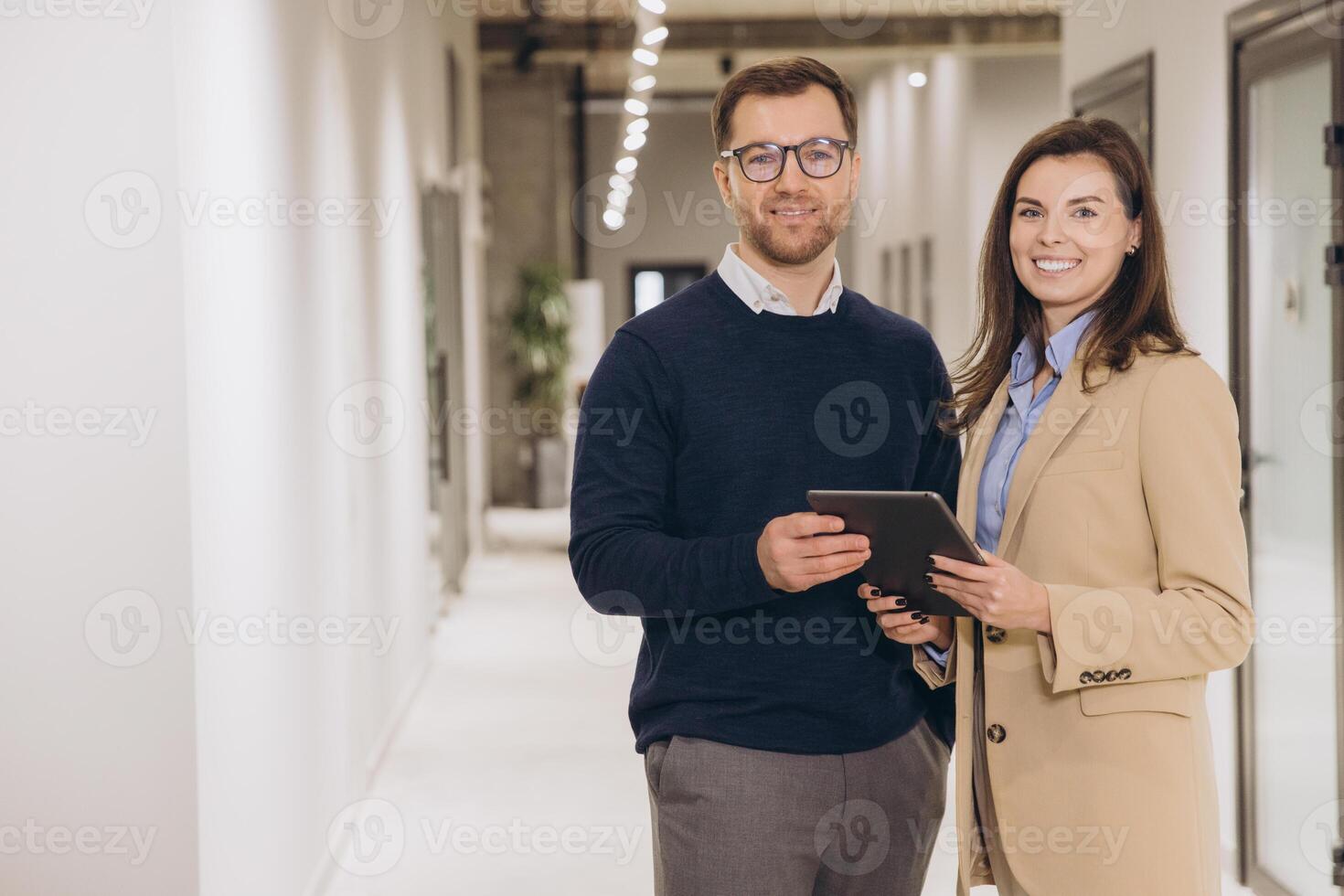 Smiling business colleagues holding a digital tablet while standing in a modern office hallway, engaging in productive discussion and collaboration photo