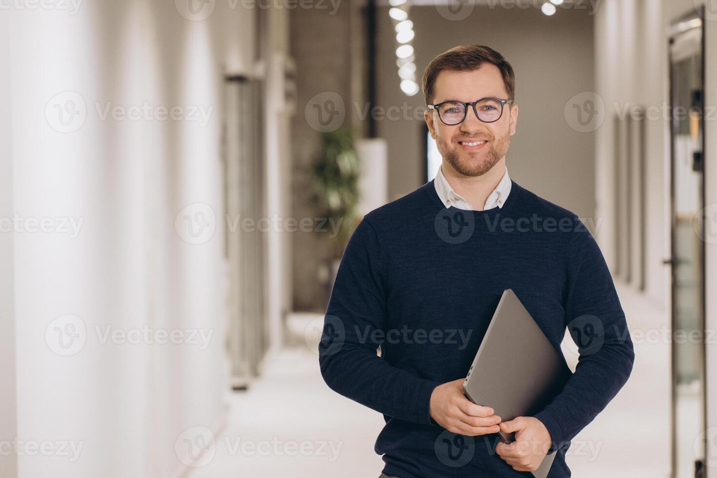 Portrait of a smiling businessman holding a laptop while walking in a bright office corridor photo