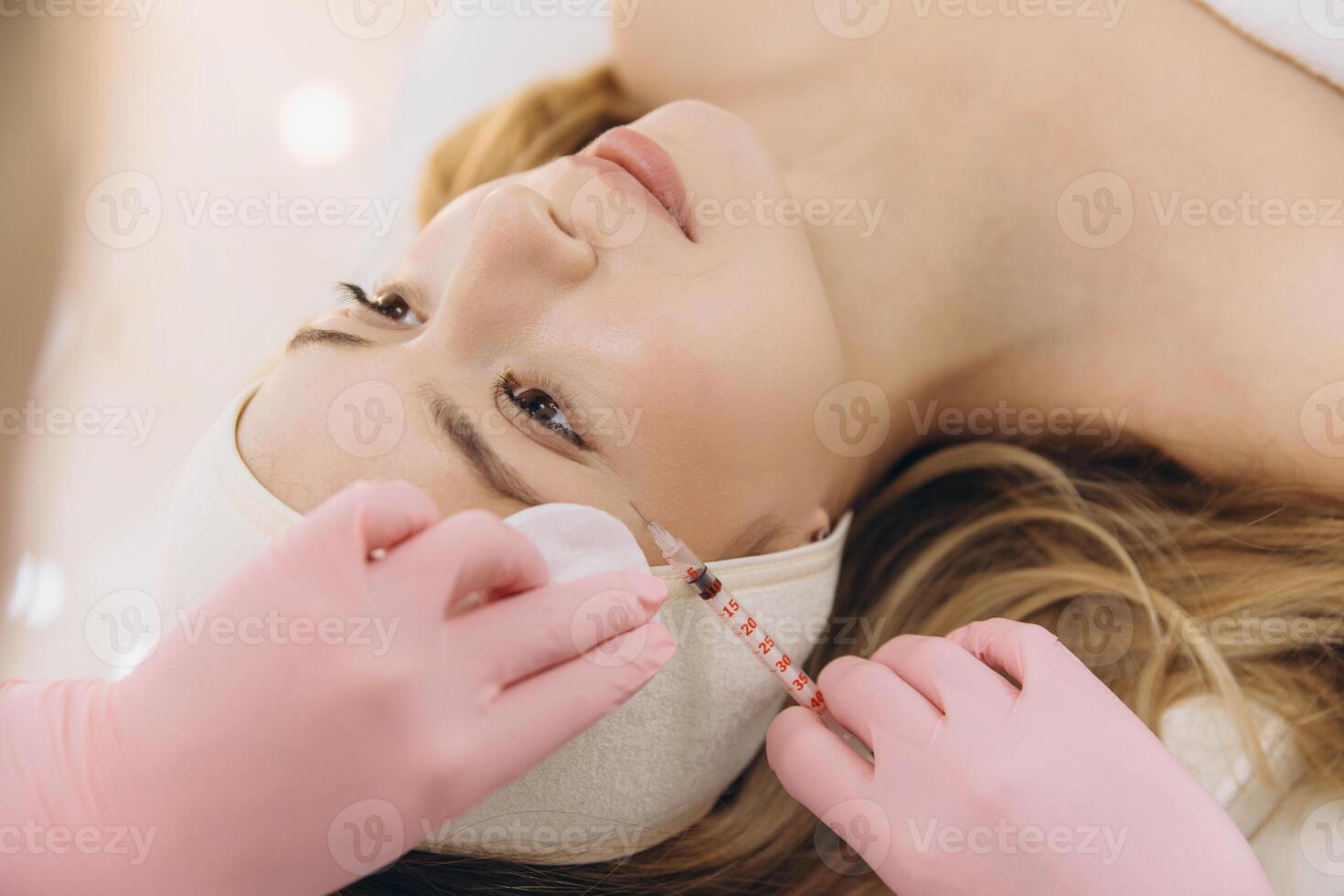 Beautician wearing pink gloves injecting botox with a syringe in forehead of young woman lying on a bed in a beauty clinic photo