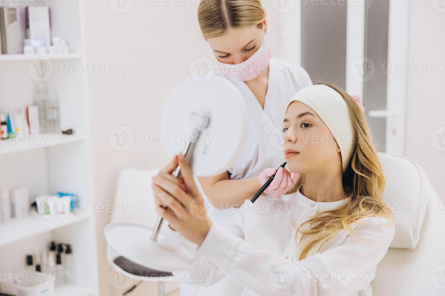 Cosmetologist marking the patient's face while she examines her reflection in the mirror, preparing for a botox injection procedure photo