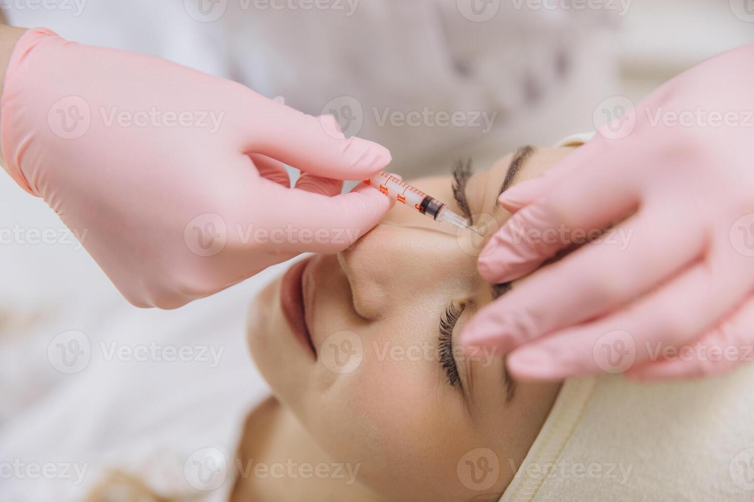 Beautician wearing pink gloves injecting botox into forehead of young woman lying on couch in beauty salon photo