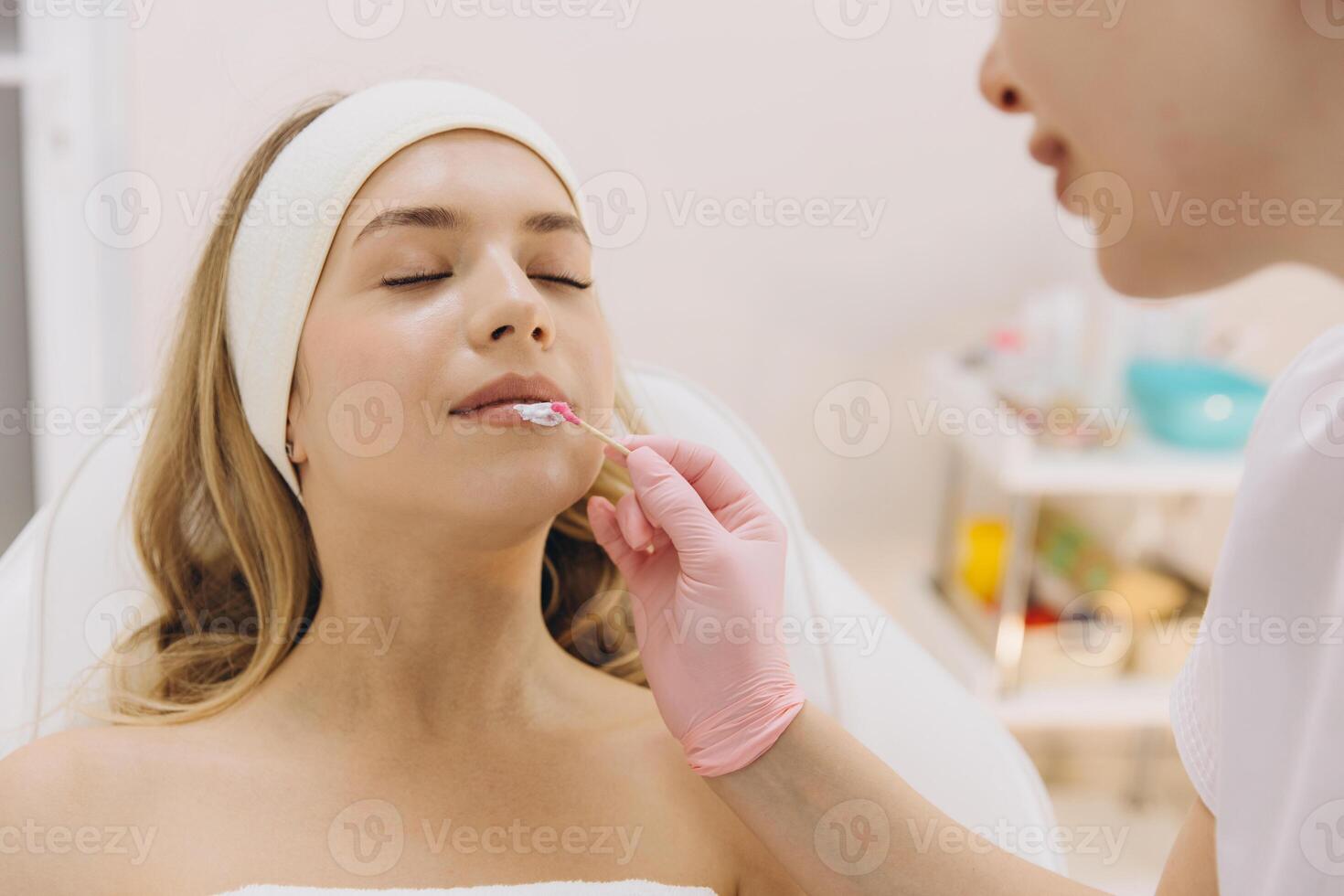 Beautician carefully applying anesthetic cream on client's lips using wooden stick, preparing for permanent makeup procedure in beauty salon photo