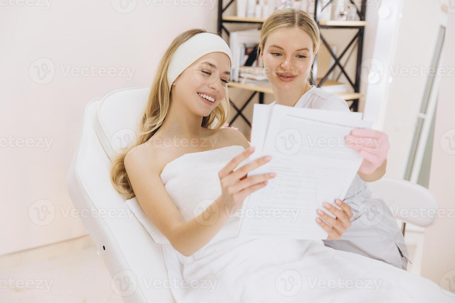 Beautician with pink gloves showing treatment plan to smiling client lying on a white chair in a beauty salon photo