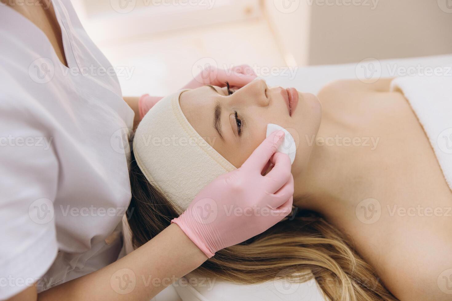 Beautician wearing pink gloves is applying skincare product on young woman's face with cotton pad photo