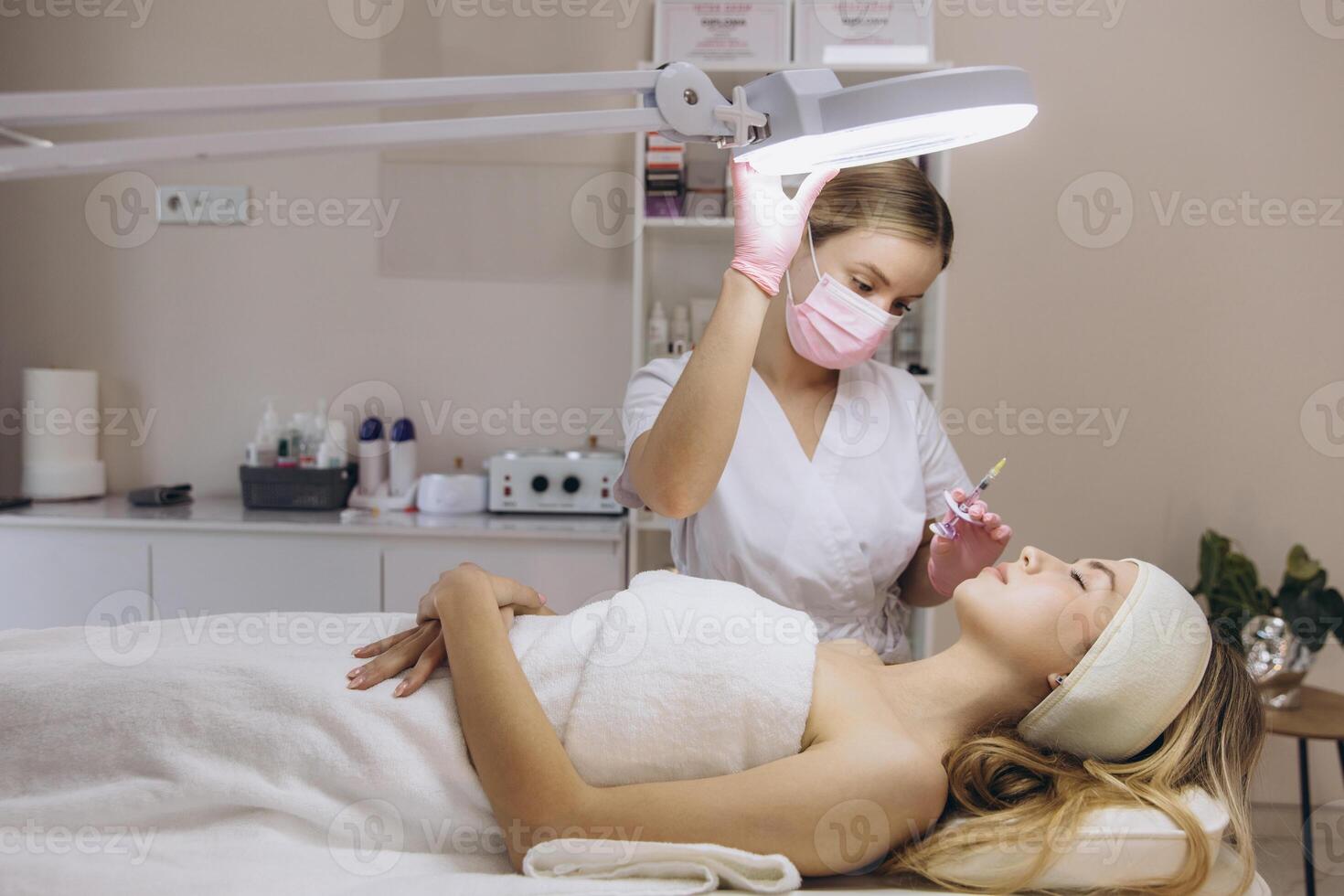 Cosmetologist adjusting lamp and giving hyaluronic acid injection to young blonde woman for facial rejuvenation in beauty salon photo