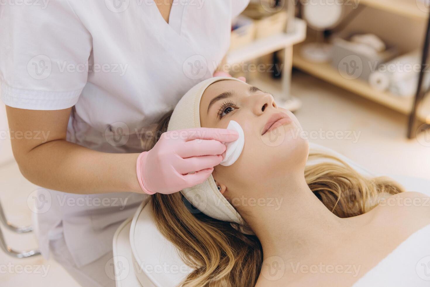 Pink gloved beautician cleansing blond client's face using cotton pad during skincare treatment photo