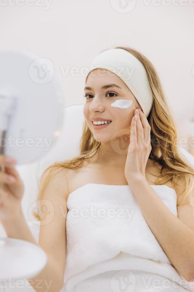 Skincare routine showing woman wearing headband, applying facial moisturizer while checking mirror during spa treatment session photo
