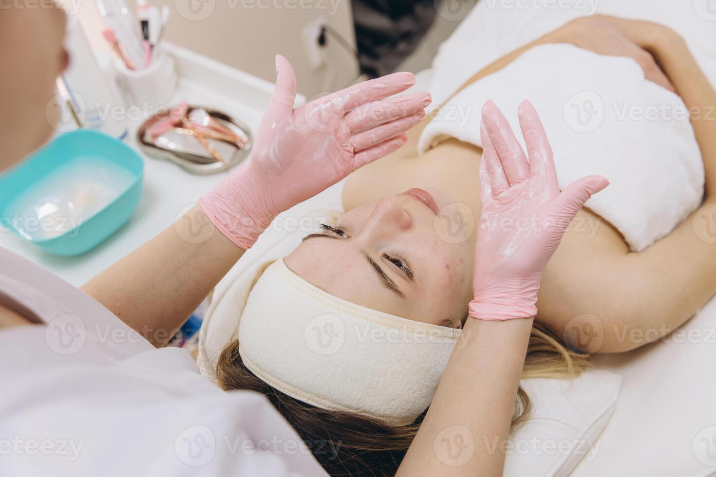 Beautician wearing pink gloves applying a facial treatment to a young woman with a headband lying on a stretcher in a beauty salon photo