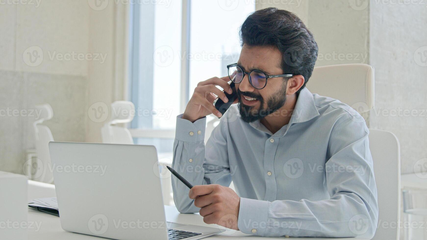 Busy multitasking Arabian businessman manager consultant Indian muslim man talk mobile phone check information on laptop computer work consulting client smartphone call business consultation in office photo