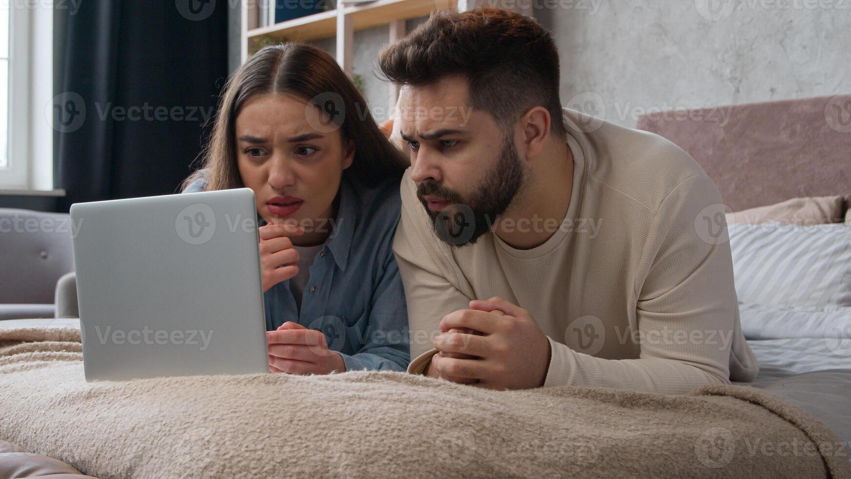 Caucasian family couple upset disappointed read bad news negative result refusal bank email upset with failure shocked woman and man homeowners on bed use laptop computer watch scary movie in bedroom photo