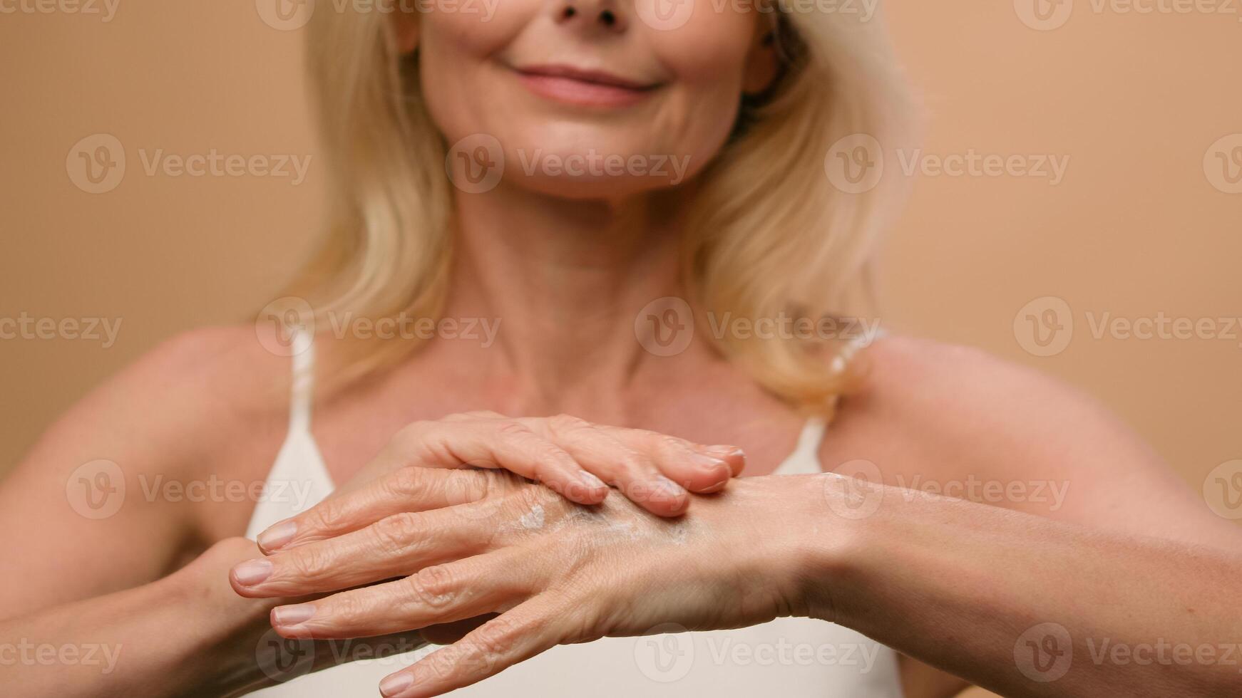 Middle-aged model mature woman apply smooth cream on hand in studio beige background female hands care rub moisturizing body lotion daily beauty treatment soft skin anti-aging cosmetics rejuvenation photo