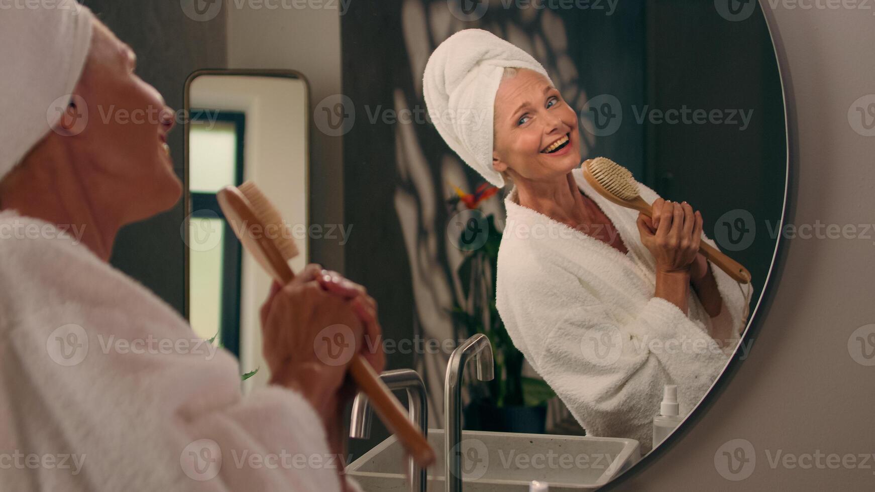 Carefree Caucasian mature senior woman in bathrobe after shower smiling middle-aged lady in home bathroom sing at hairbrush microphone dance point finger at bath mirror singing moving to music vocal photo