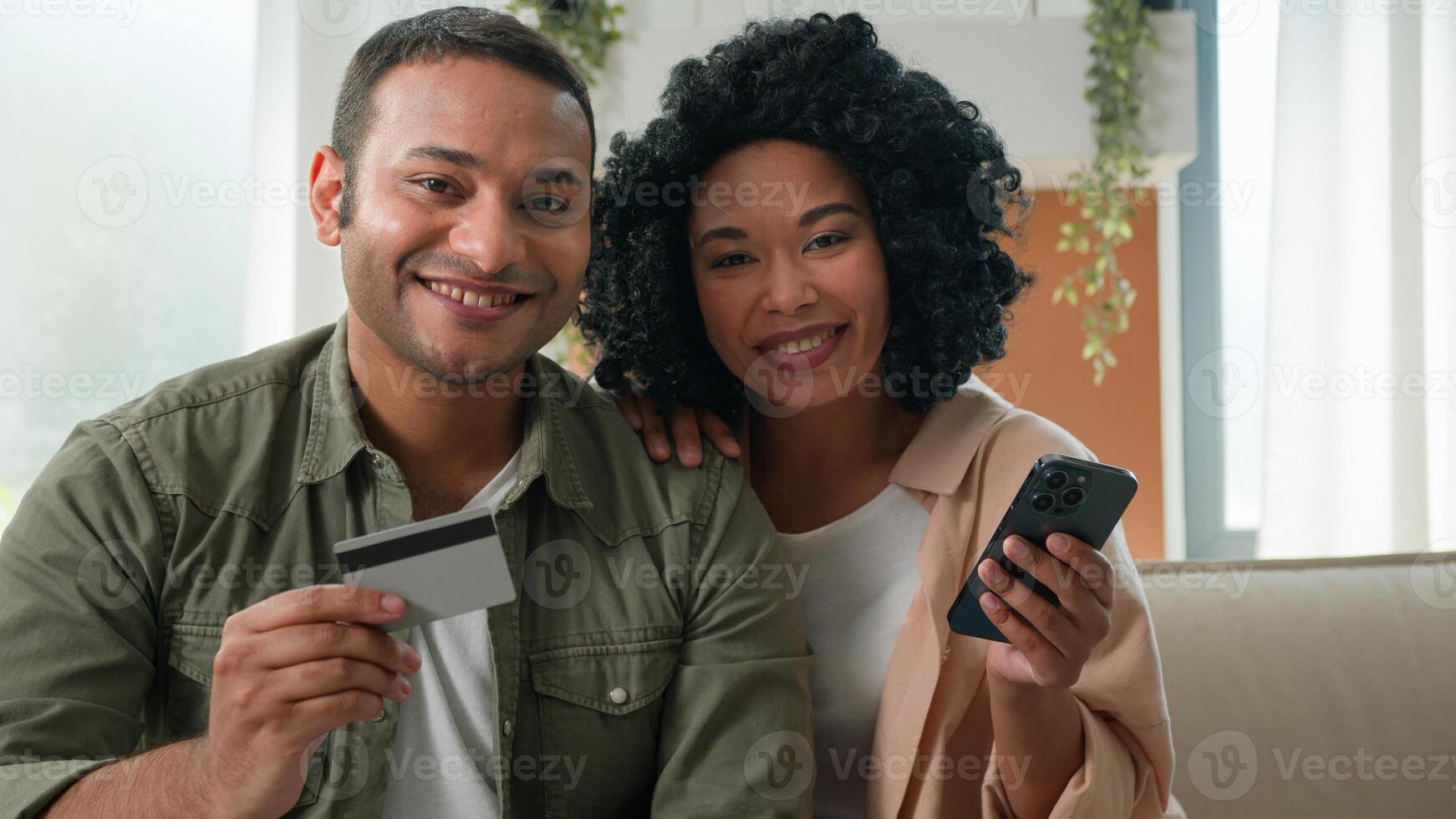 Multiracial African American Arabian family hold mobile phone and bank credit card online shopping couple clients woman and man booking delivery at home paying money budget happy look at camera smile photo