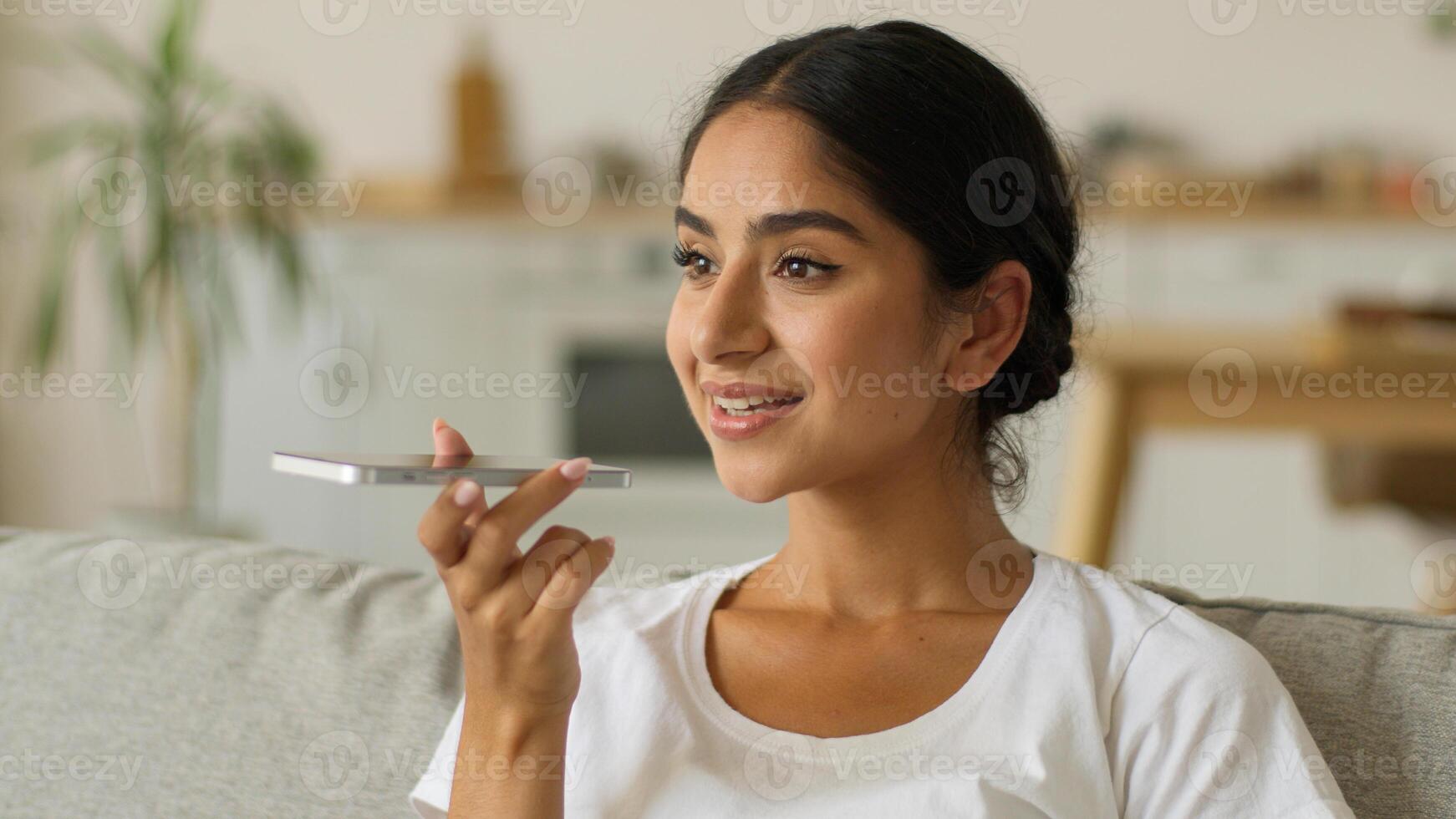 Happy smiling Indian woman at home Arabian positive Arabian girl female lady at living room mobile phone talking microphone smartphone mic talk loudspeaker record voice mail recording audio message photo