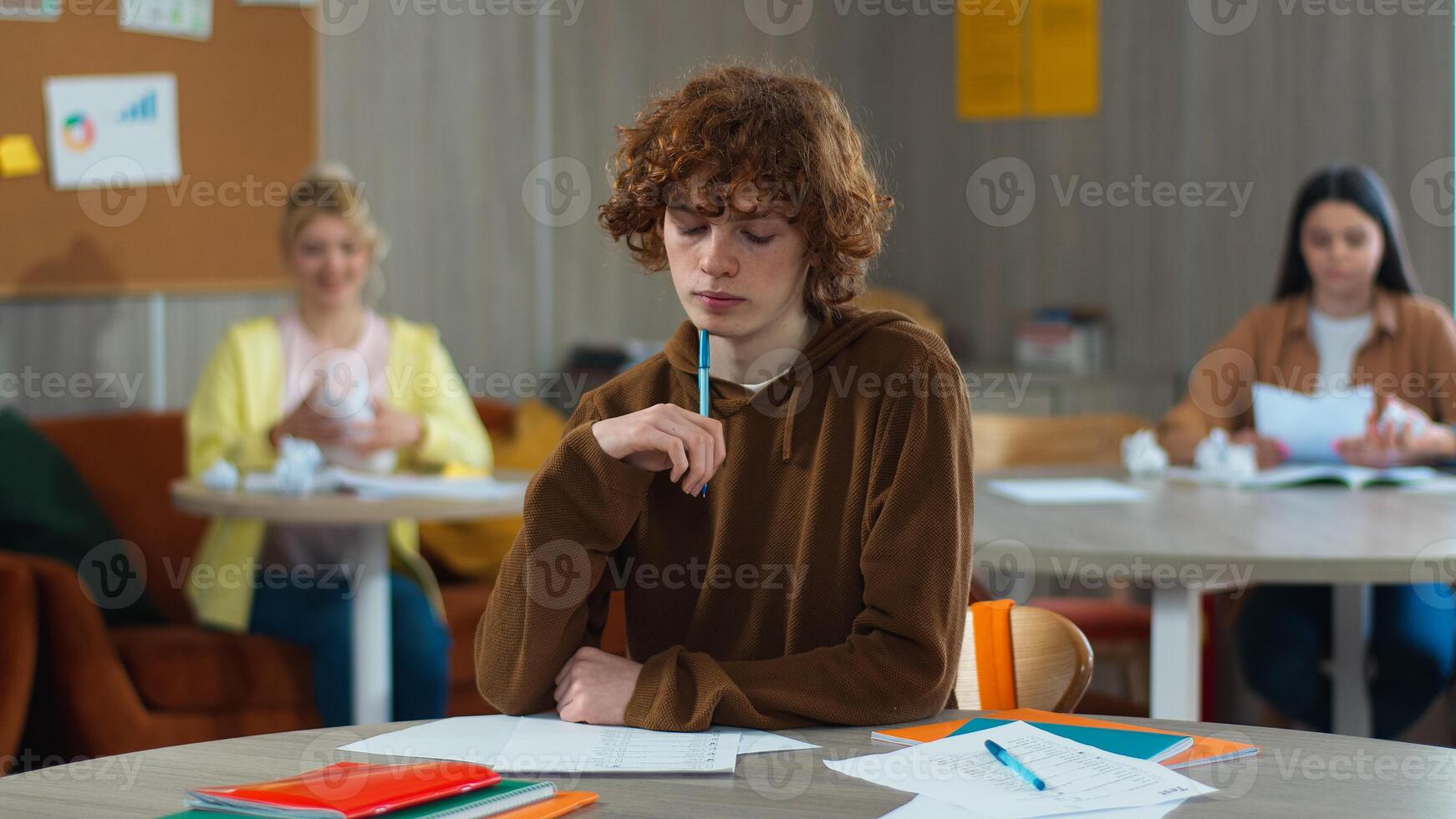 High school bullying upset stressed lonely Caucasian guy teen male boy schoolboy nerd student teenager writing task class desk university college children students mock bully throwing papers balls photo
