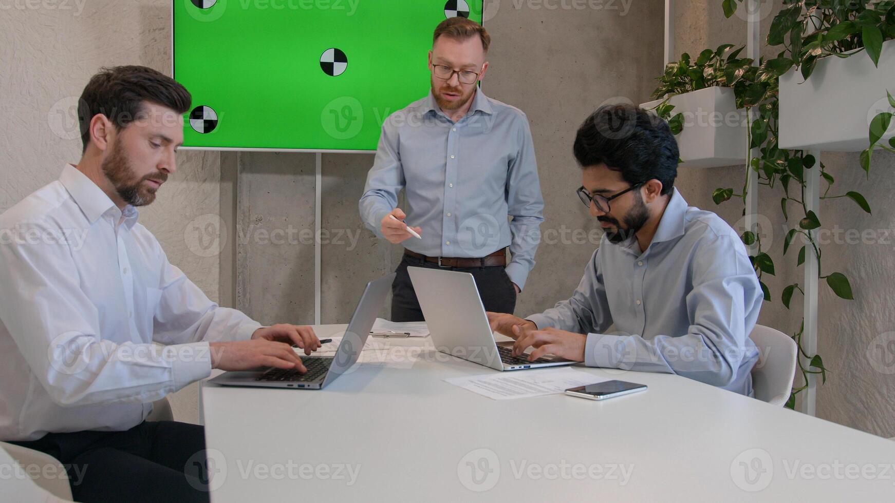 Male leader speaker coach talking to team multiracial men listen to CEO businessman teaching employees showing business presentation at group conference pointing at green interactive board chroma key photo