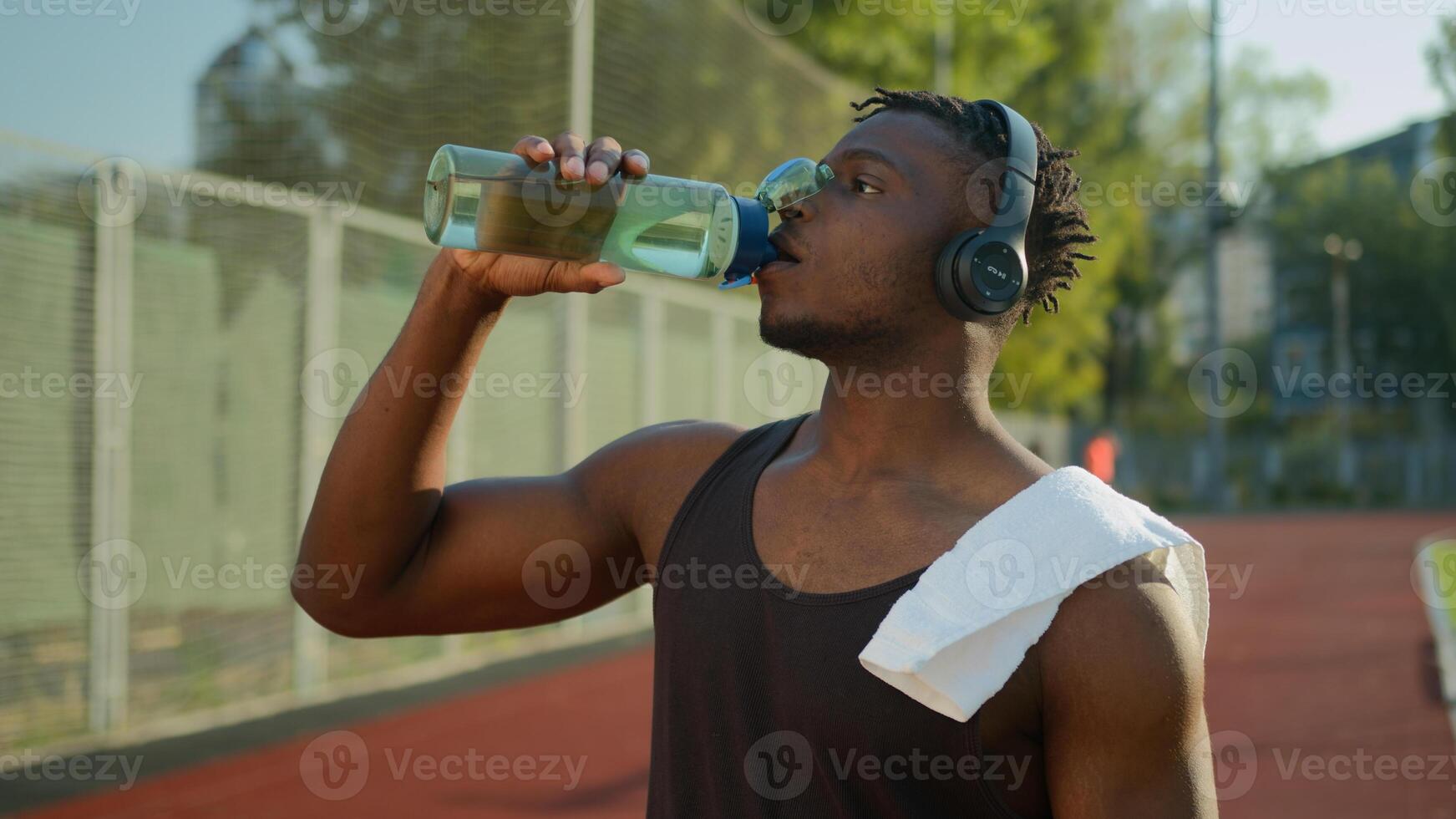 Tired African American sport man male muscular sportsman runner athlete drinking water sports bottle refresh body hydration after running on stadium workout outdoors drink listen music in headphones photo