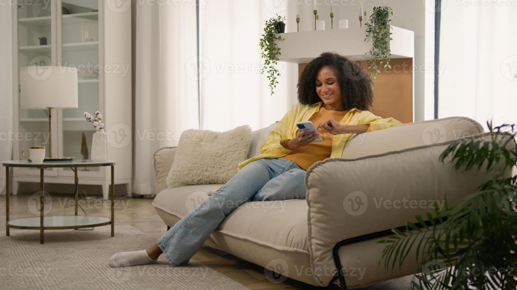 African American girl smiling young woman holding mobile phone using modern device on couch at home scrolling smartphone read social network laughing female lady blogger gadget addicted buying online photo