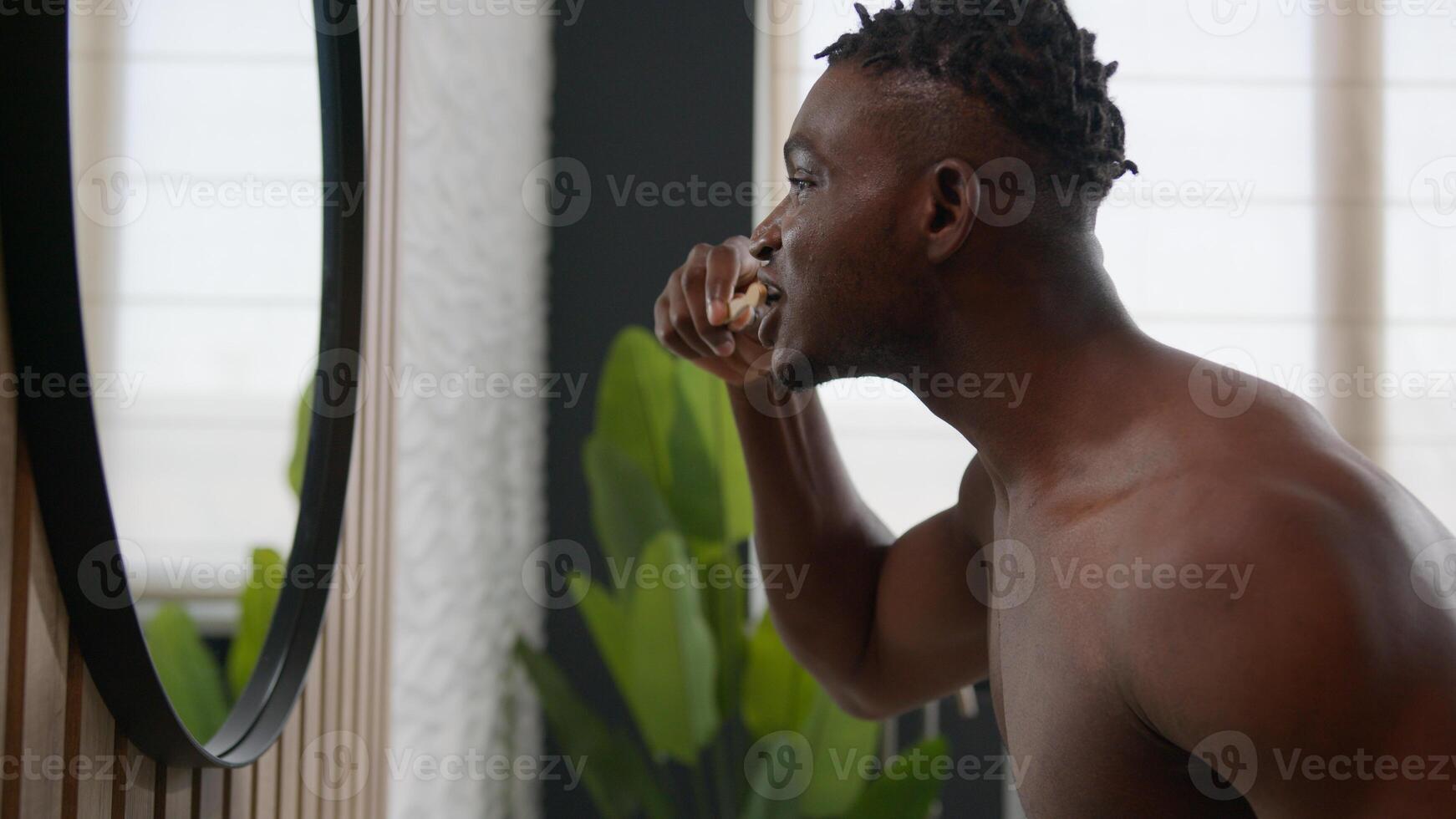 African American ethnic man in bathroom brushing teeth with toothbrush toothpaste looking at bath mirror cleaning gums tooth plaque oral care morning dental healthy hygiene procedure caries protection photo
