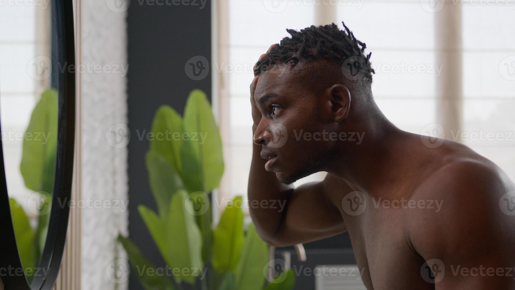 African American man looking in mirror touch coarse hair dreadlocks upset anxious guy suffer baldness alopecia problem skincare treatment with hairstyle repair cosmetic product male beauty in bathroom photo