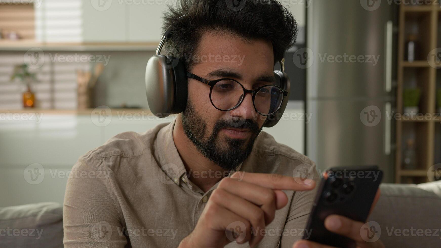 Happy Arabian Indian man in headphones listen music at home kitchen male millennial rest on couch choose song musical track in playlist online audio service on mobile phone enjoying high quality sound photo