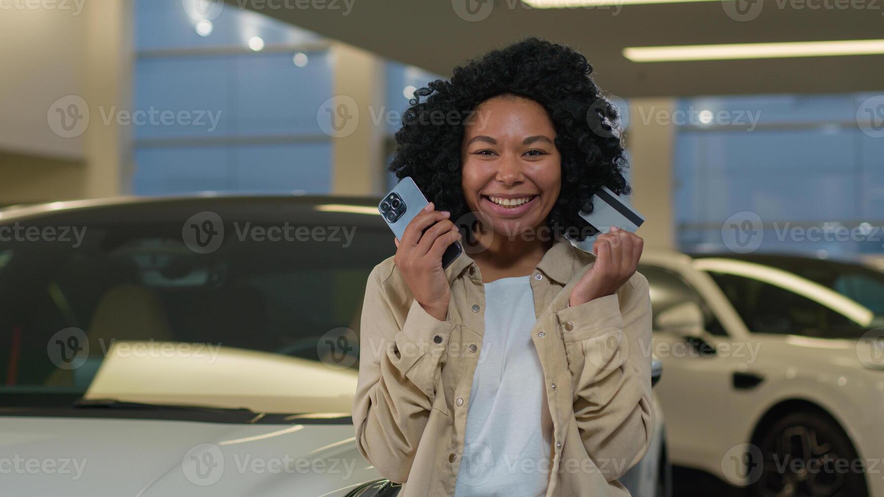 African American happy customer woman buyer hold credit card mobile phone shopping buying new luxury electric automobile in dealership auto salon purchase vehicle credit payment car sharing service photo