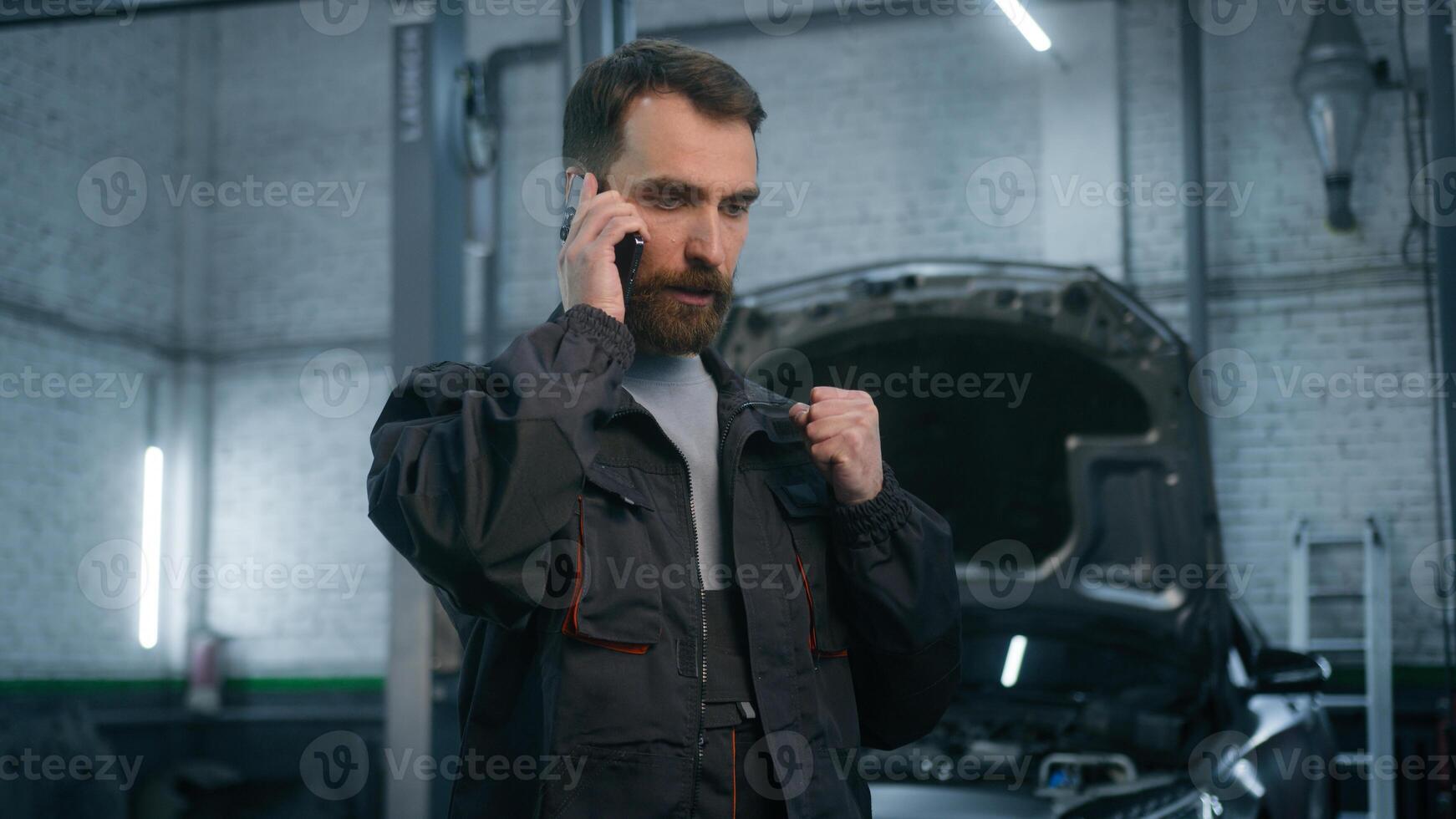 Annoyed serious Caucasian adult 30s man in uniform auto mechanic engineer talking mobile phone in repair car service male worker of technical industry communicate speak smartphone at vehicle workshop photo