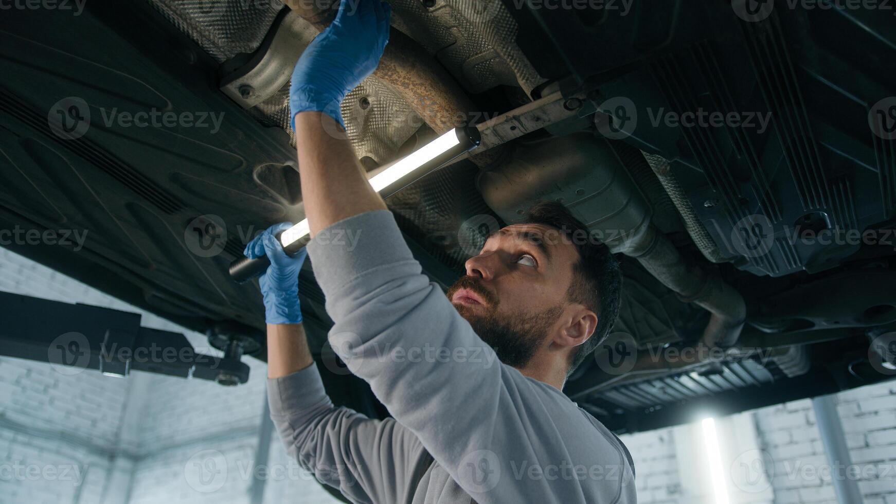 Caucasian guy mechanical serviceman working underneath car in garage checking automobile maintenance mechanic man in auto repair service shop vehicle diagnostics survey truck using flashlight light photo