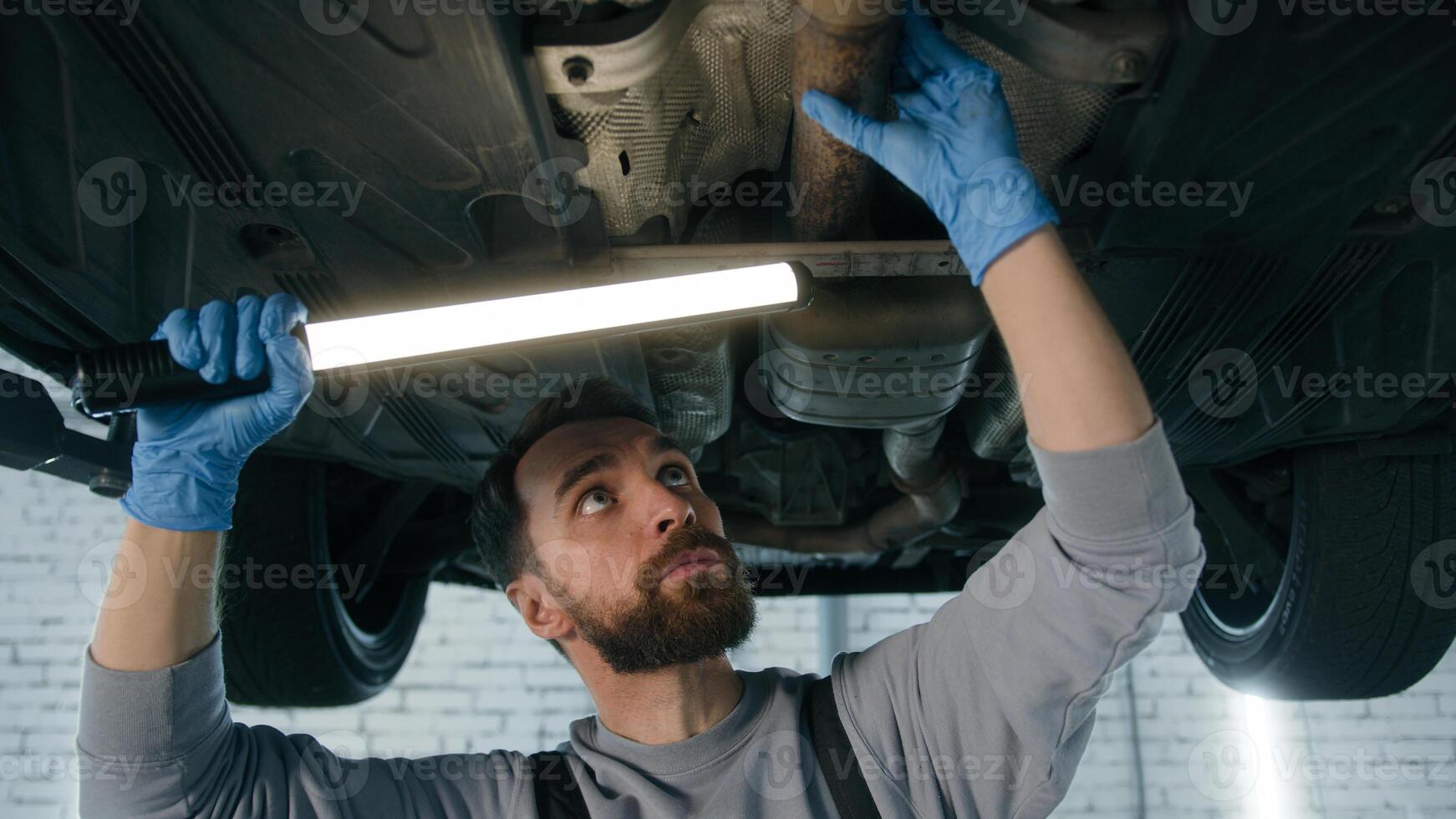 Caucasian man guy repairman auto mechanic working underneath lifting vehicle at garage station checking automobile steel details suv truck maintenance using work illuminate light at repair car service photo