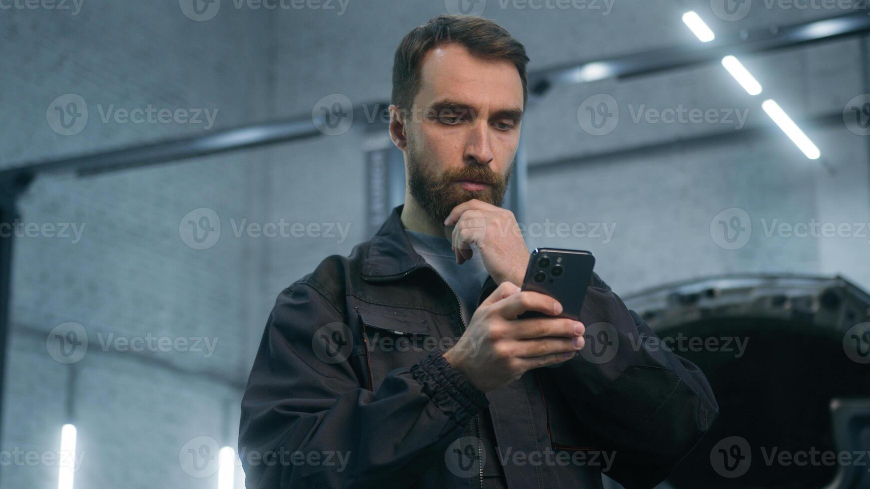 Thoughtful pensive guy Caucasian man mechanic car serviceman using smartphone thinking make online order internet store in auto service automobile business male factory worker looking at mobile phone photo