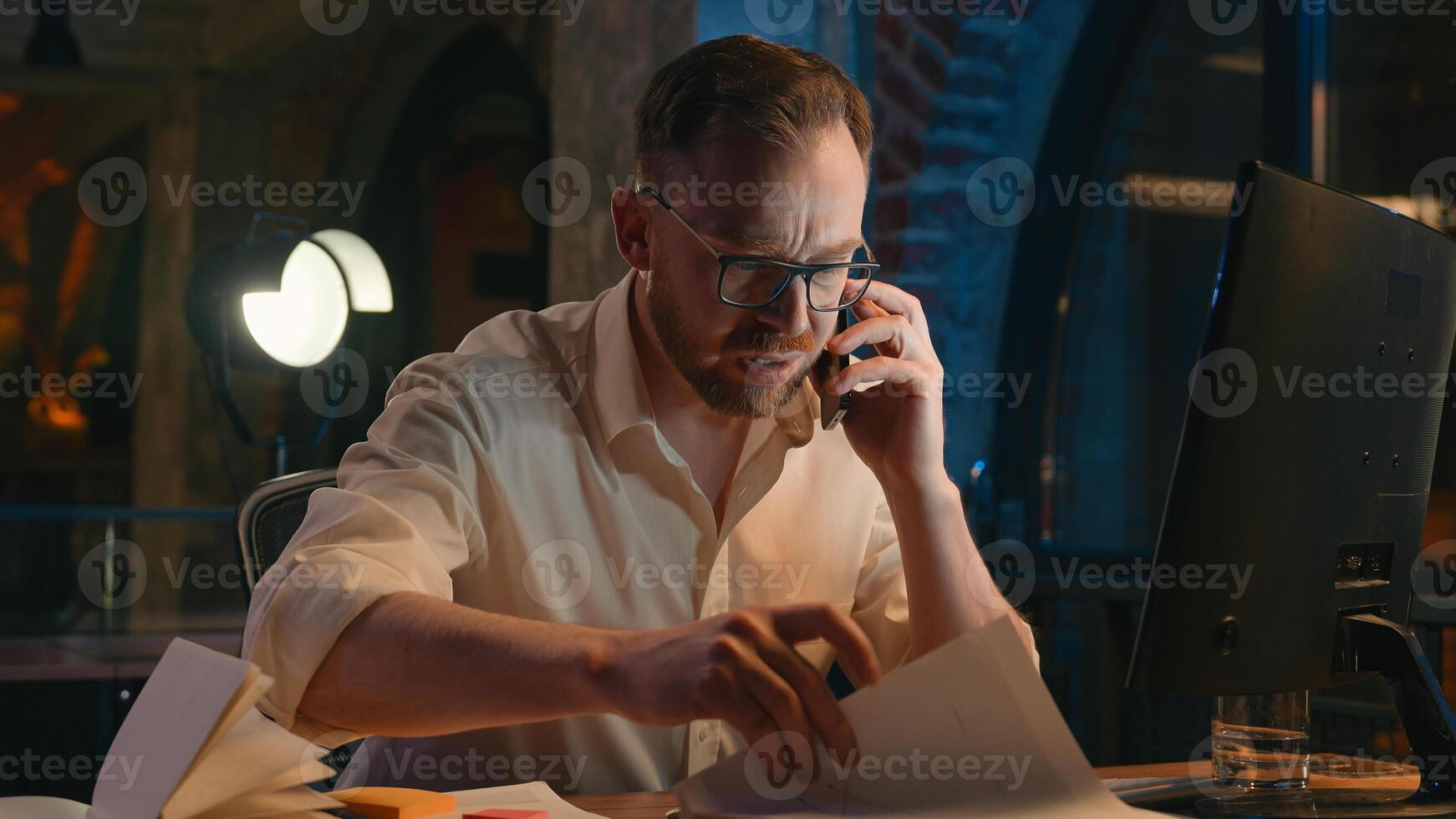 Caucasian man business guy businessman male in dark night evening office work calling mobile phone call talking dissatisfied sad upset confused smartphone searching papers documents paperwork deadline photo