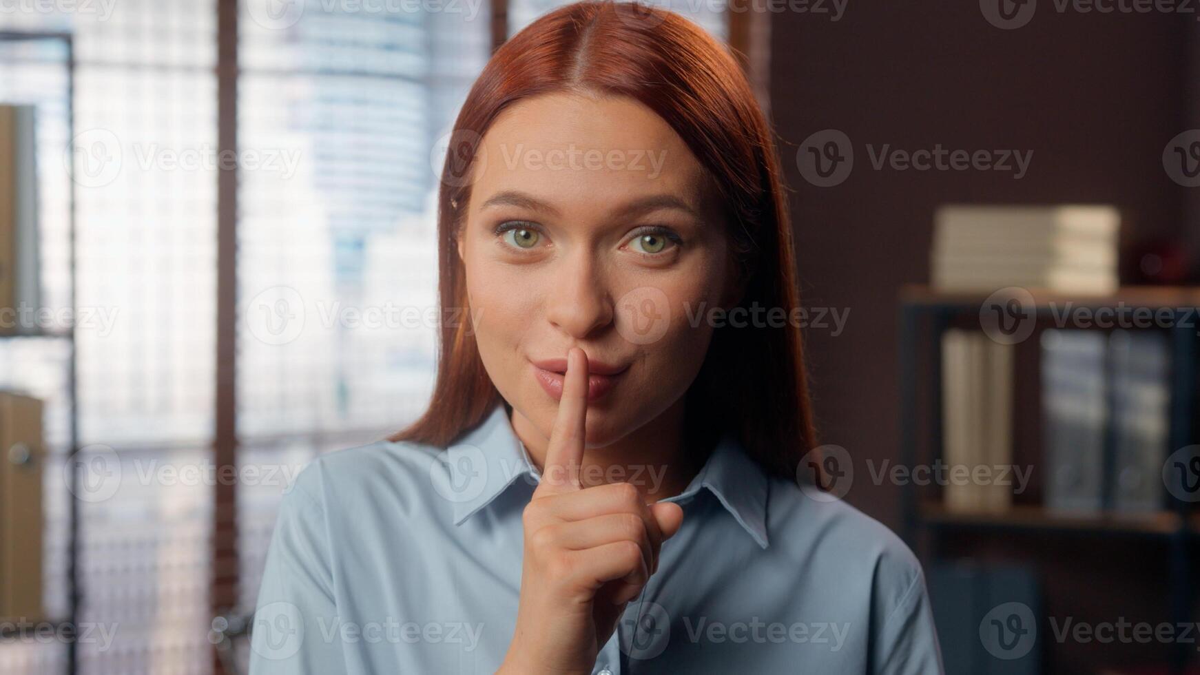 Smiling Caucasian business woman girl female businesswoman employer manager in office hold index finger near lips mouth hush silence quiet gesture secret secrecy information silent looking at camera photo