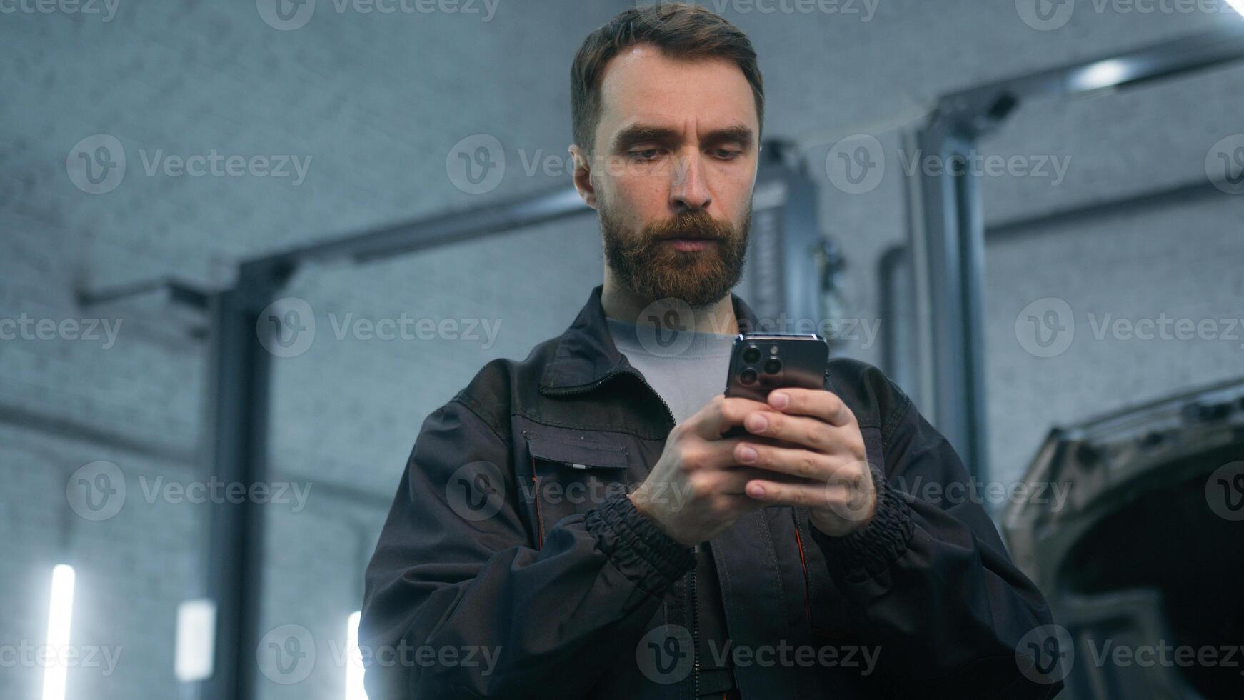 Mechanic man auto mechanical worker using mobile phone gadget in repair car service garage automobile factory pensive thoughtful male guy repairman look at smartphone screen think typing on cellphone photo