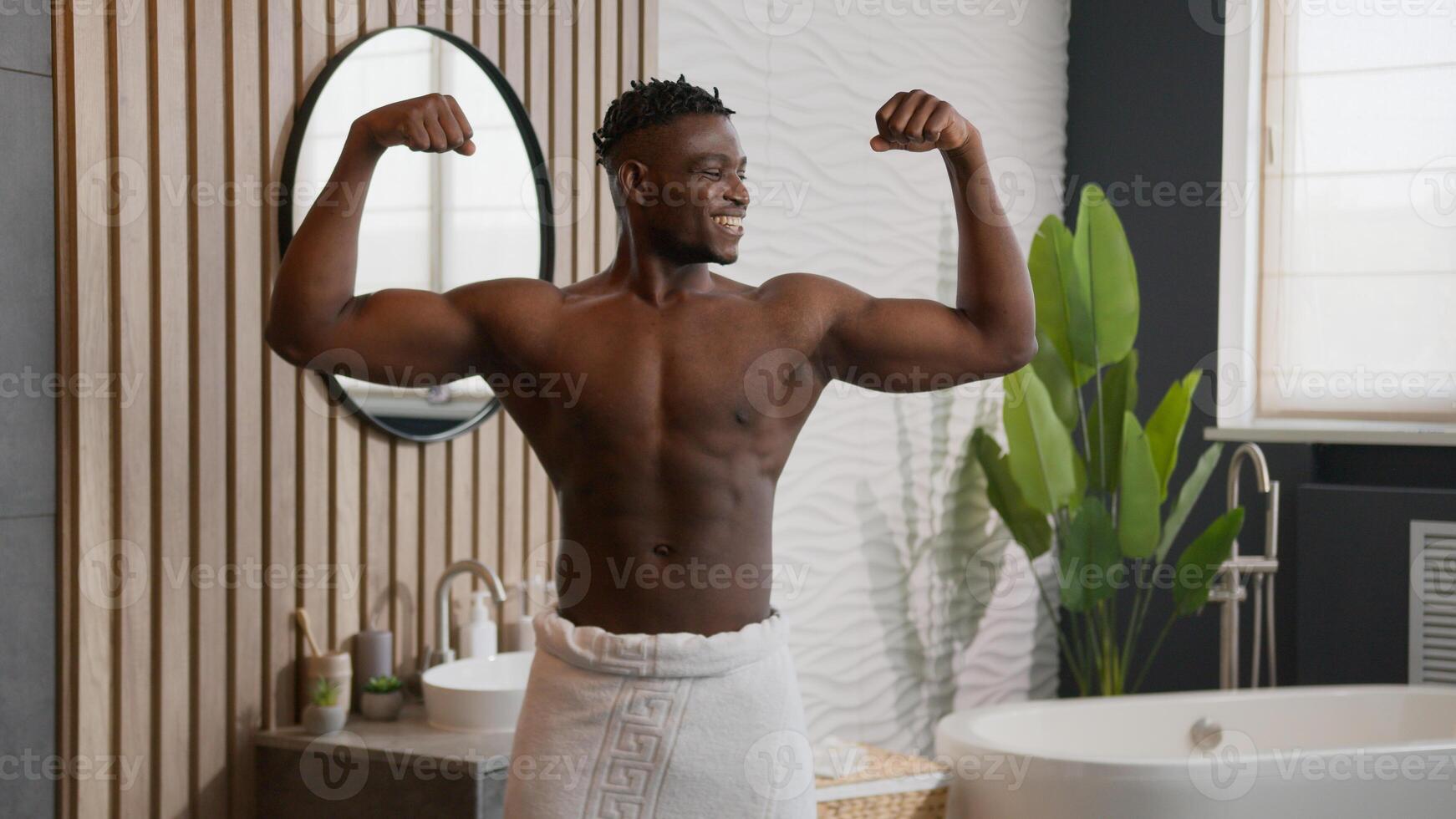 Muscular African American man smiling athletic male bodybuilder with bath towel on hips showing muscles biceps raising arms sport strong power sexy ethnic guy naked torso in bathroom care of skin body photo