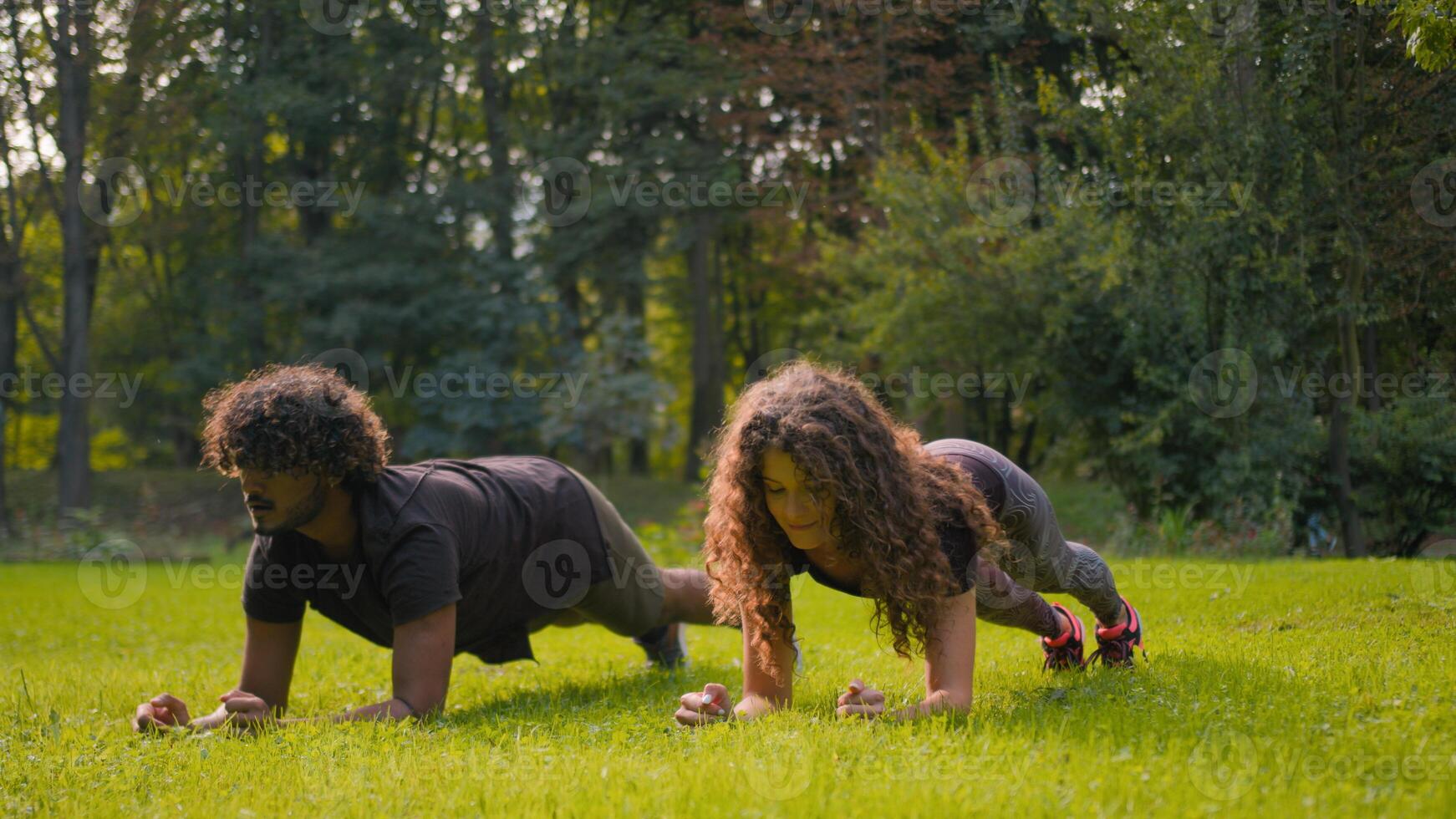 Fitness couple diverse Arabian man Caucasian woman doing sport morning practice city park nature outside workout sporty strong sportsman sportswoman bodybuilders plank exercise on grass lawn outdoors photo