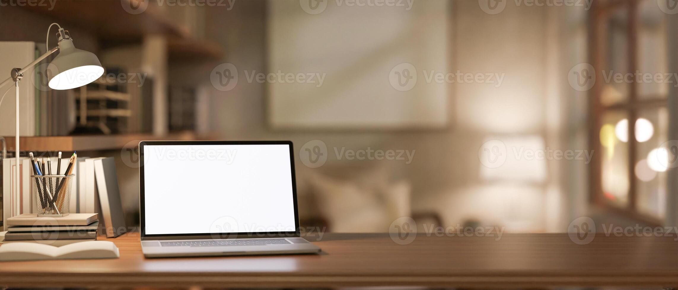 A close-up of a laptop and stationery on a wooden desk, illuminated by a table lamp. photo