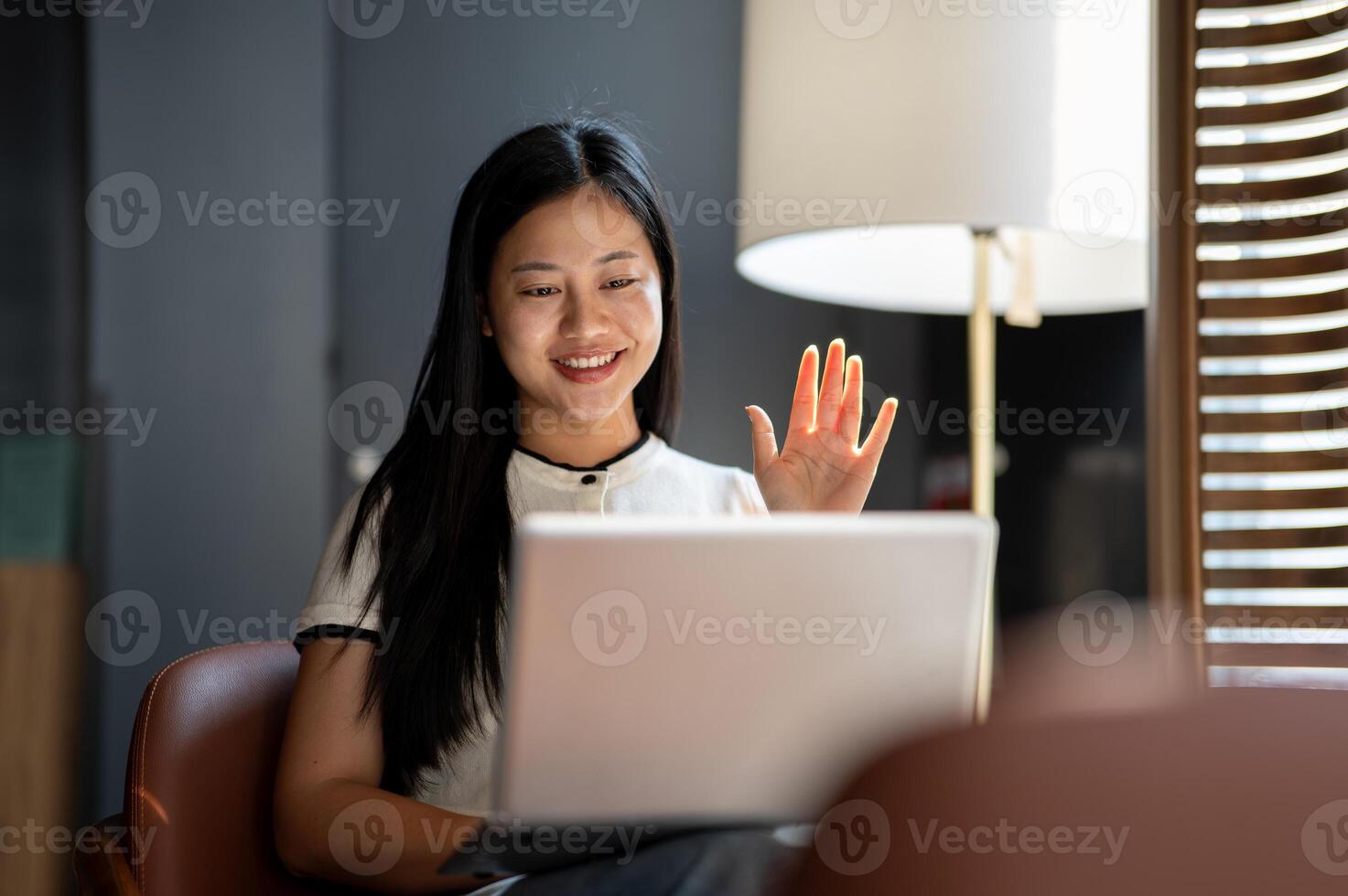 A smiling Asian woman has an online meeting with her team while working remotely at a coffee shop. photo