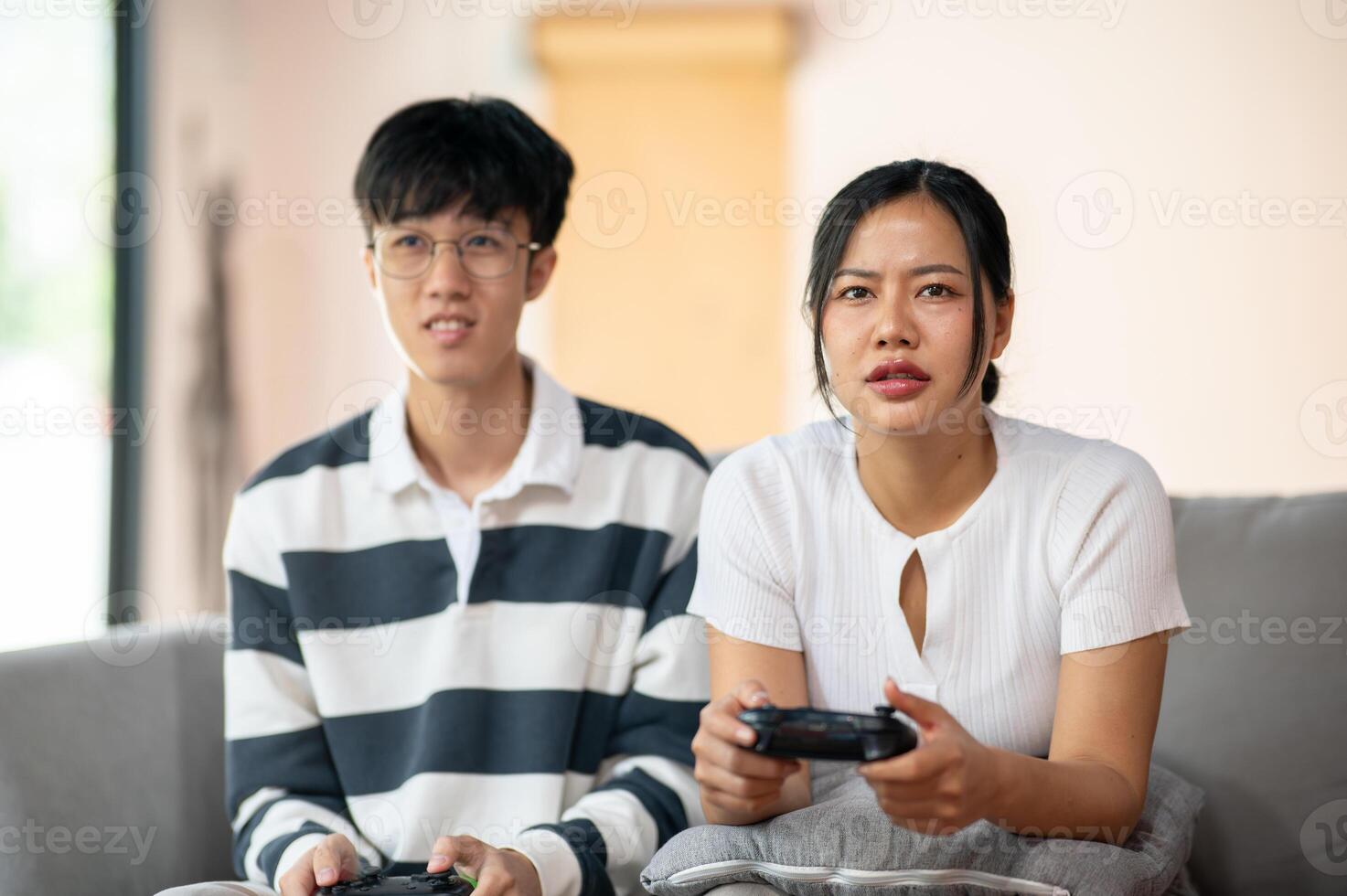 Two Asian friends excitedly play a game with joysticks in the living room, having a fun time. photo