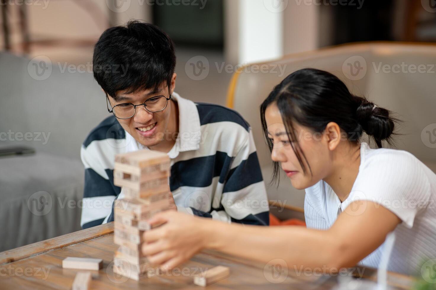Two cheerful Asian friends enjoy playing Jenga, focusing on removing a wooden block. photo