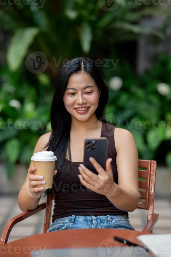 A happy Asian woman reads a chat on her smartphone while enjoying coffee at an outdoor table. photo