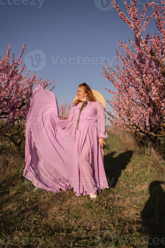 Woman blooming peach orchard. Against the backdrop of a picturesque peach orchard, a woman in a long pink dress and hat enjoys a peaceful walk in the park, surrounded by the beauty of nature. photo